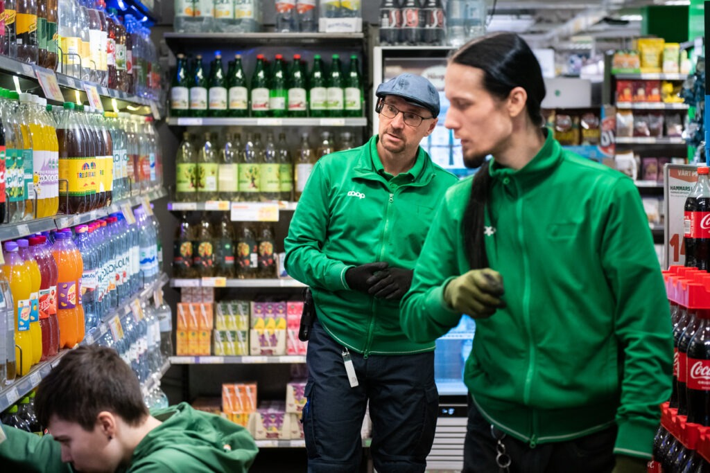 Tre män i gröna Coop-kläder vid en hylla inne i butiken. En av männen arbetar på huk på golvet.