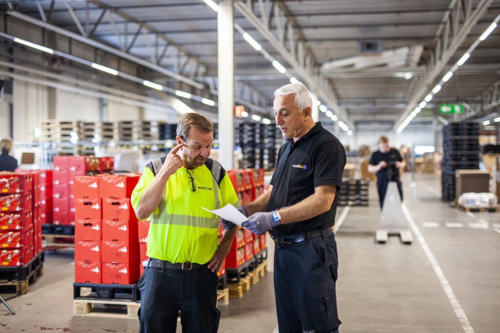 Två män i arbetskläder står inne på ett lager. De diskuterar ett papper som den ena håller i handen.