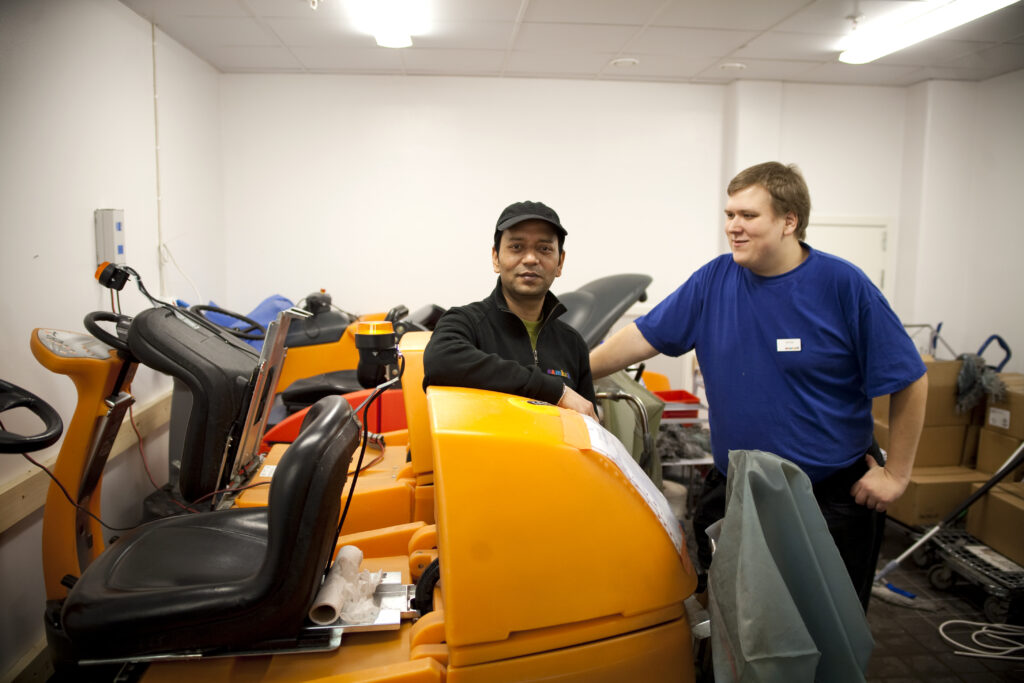 Två män står vid arbetsredskap. En av dem, med svart fleecejacka och svart keps, tittar in i kameran.