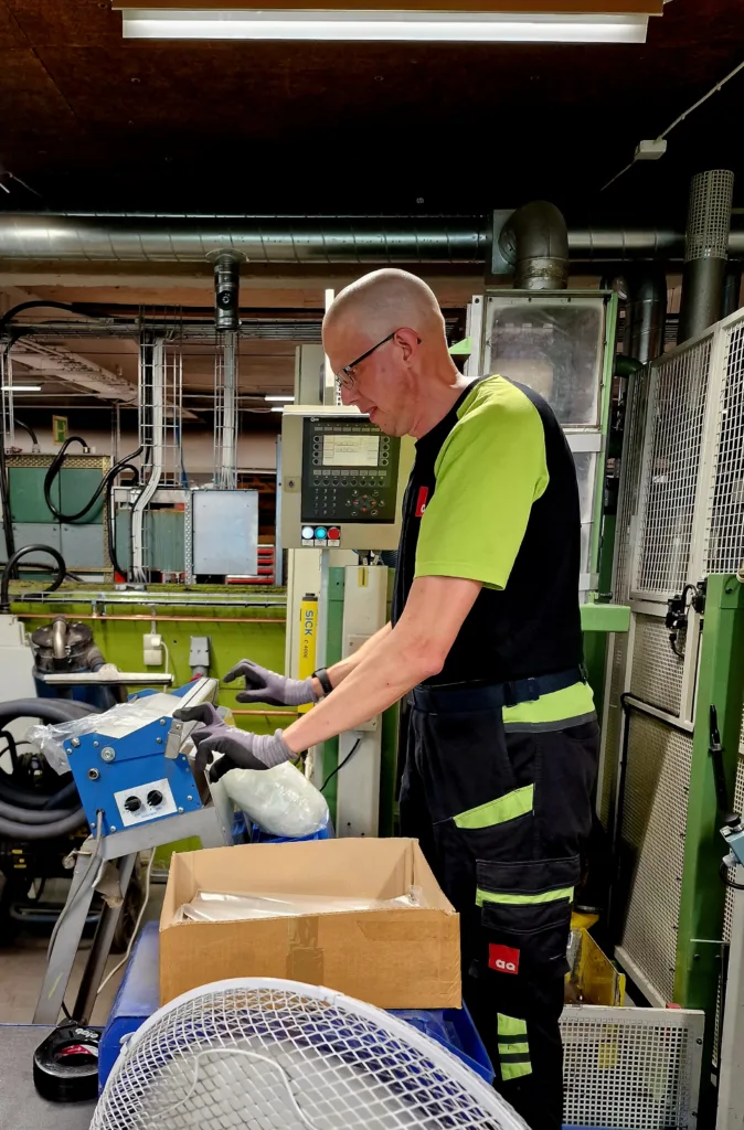 Fredrik står vid sin maskin och arbetar. Han är fokuserad på arbetsuppgiften och tittar inte in kameran.
Han bär arbetshandskar och en gul och svart t-shirt.