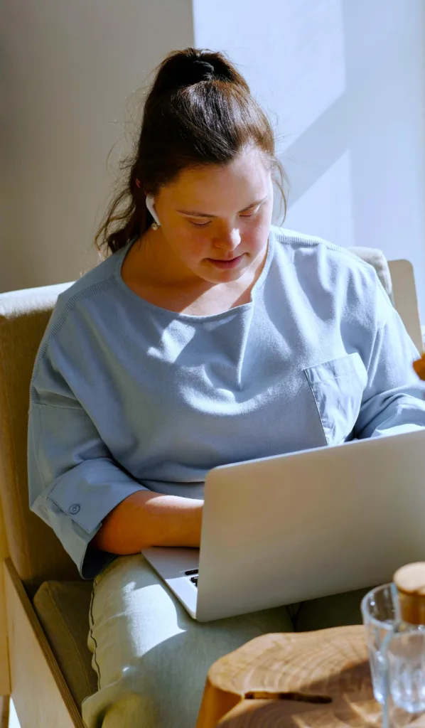 Tjej sitter och skriver med datorn i knät. Hon har Downs. Hon har mörkt hår uppsatt i en hög hästsvans och hon bär en ljusblå tröja.