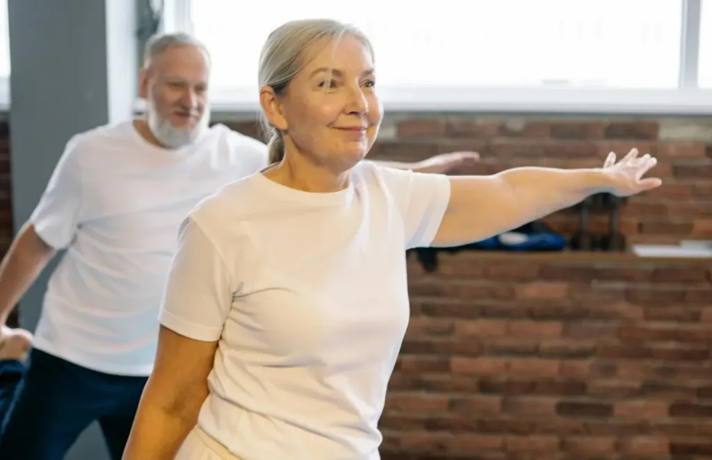 En kvinna och en man inne i en träningslokal. De balanserar på ett ben, stretchar lårmusklerna och sträcker samtidigt ut vänsterarmen.