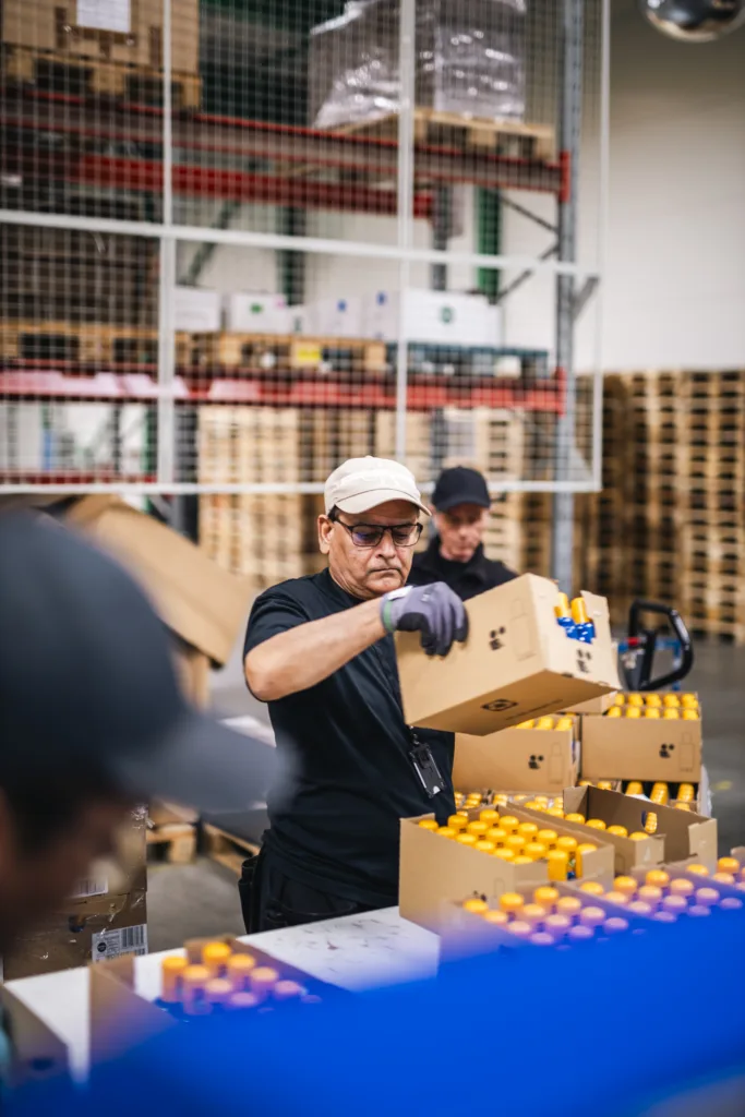 En man står och arbetar på ett lager. Han bär vit keps, glasögon och grå arbetshandskar.