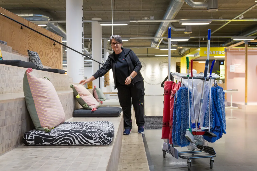 Bild inifrån en butik. En servicevärd, kvinna, står och rättar till kuddar vid en sittbänk.