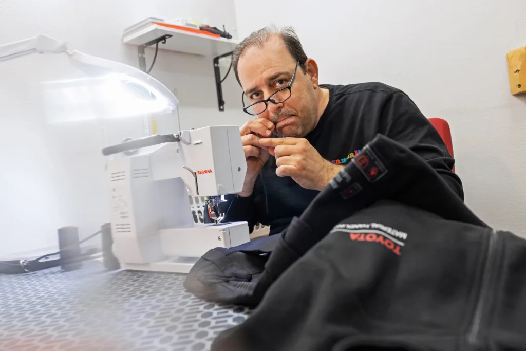 En man i glasögon sitter vid en symaskin. Det ser ut som att han har en sytråd i munnen och just ska trä symaskinen.