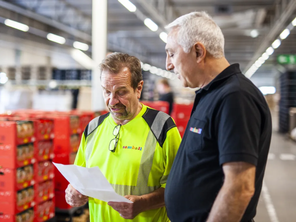 Två män i Samhallkläder står inne på ett lager och pratar med varandra. Den ena mannen håller i ett papper som de ser ut att diskutera.