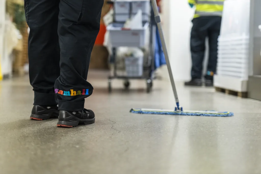 Någon städar ett golv. Man ser svarta arbetsbyxor med Samhall-logga, svarta skor och en blå mopp.