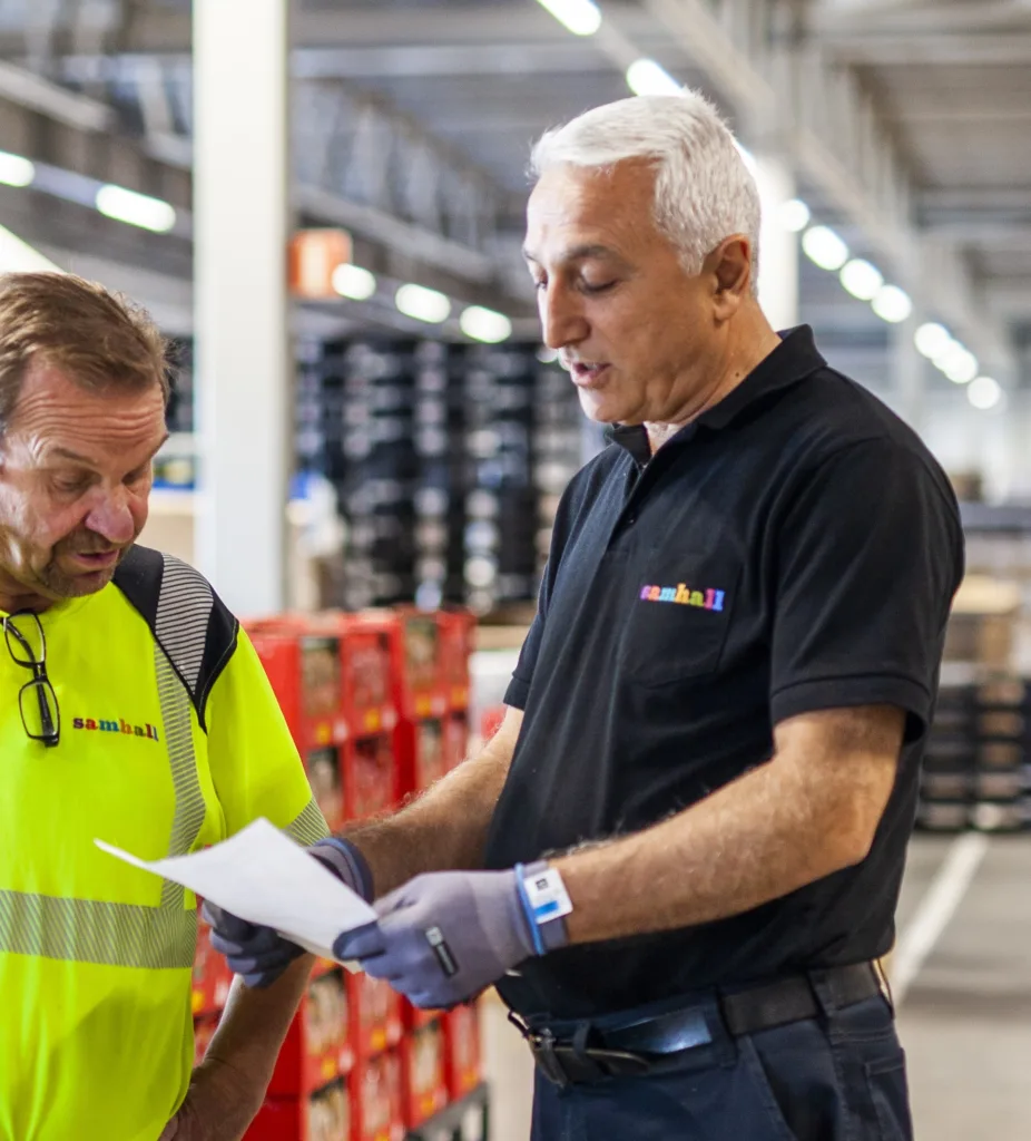 Två män i Samhallkläder står och pratar. En av dem håller i ett papper.