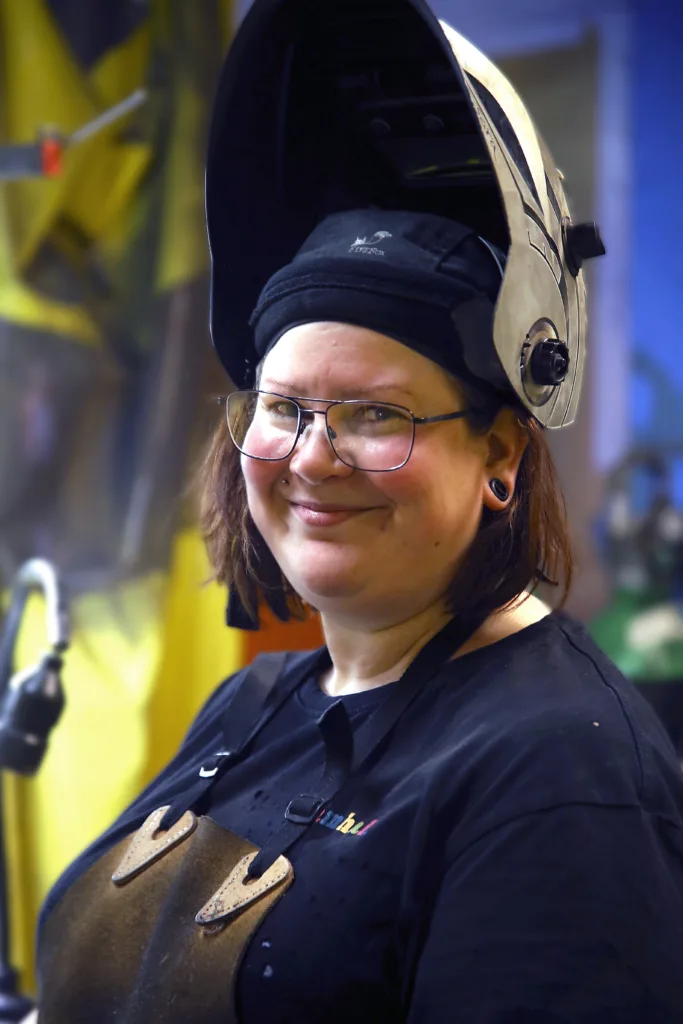 Svetsaren Sara har fällt upp visiret på sin svetshjälm. Hon ler och tittar in i kameran.
