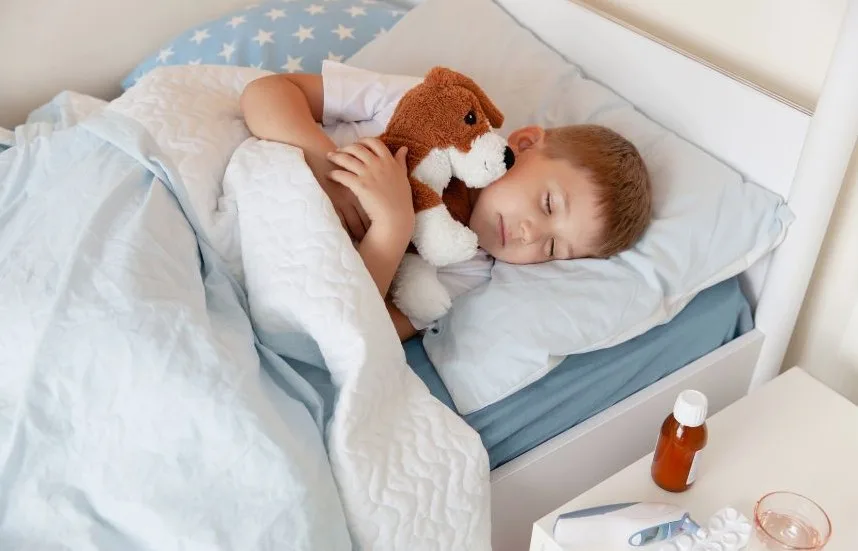 Liten pojke i sin säng. Han sover och kramar om en vit och brun gosedjurshund. På sängbordet står en medicinflaska.