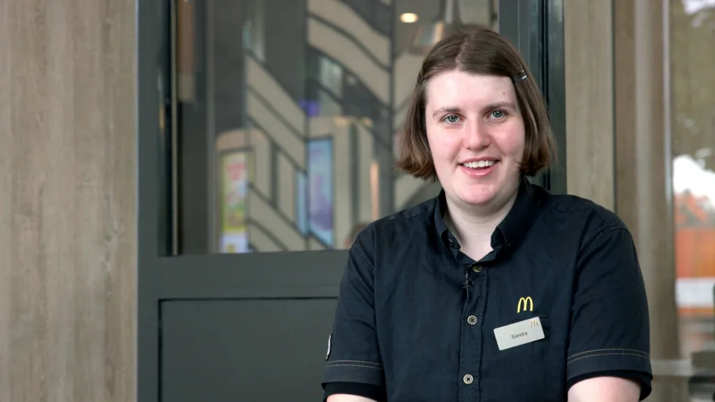Sandra ler och tittar in i kameran. Hon bär en namnskylt och en svart tröja som det står McDonalds på.