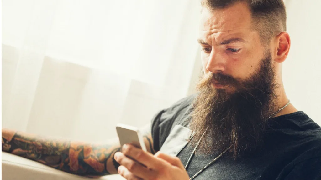 Man med långt skägg, svart T-shirt och tatuerade underarmar. Han sitter och läser i mobilen.