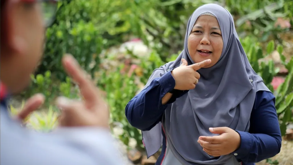 Kvinna använder teckenspråk. Hon bär en gråblöja huvudduk och långärmad, mörkblå tröja. Hon står utomhus med mycket grönska bakom sig.