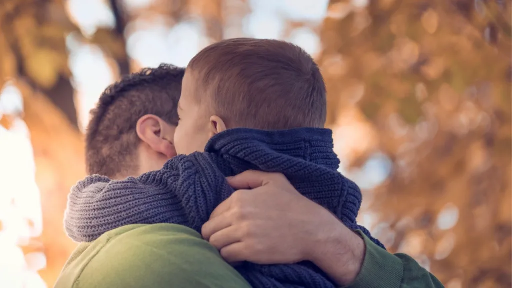 En liten pojke i blå tröja blir buren och omfamnad av en man. Pojken har sina armar runt mannens nacke.