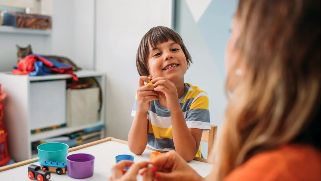 Pojke i 6-årsåldern sitter vid ett bord och leker. Man ser ryggen av en vuxen kvinna vid samma bord.