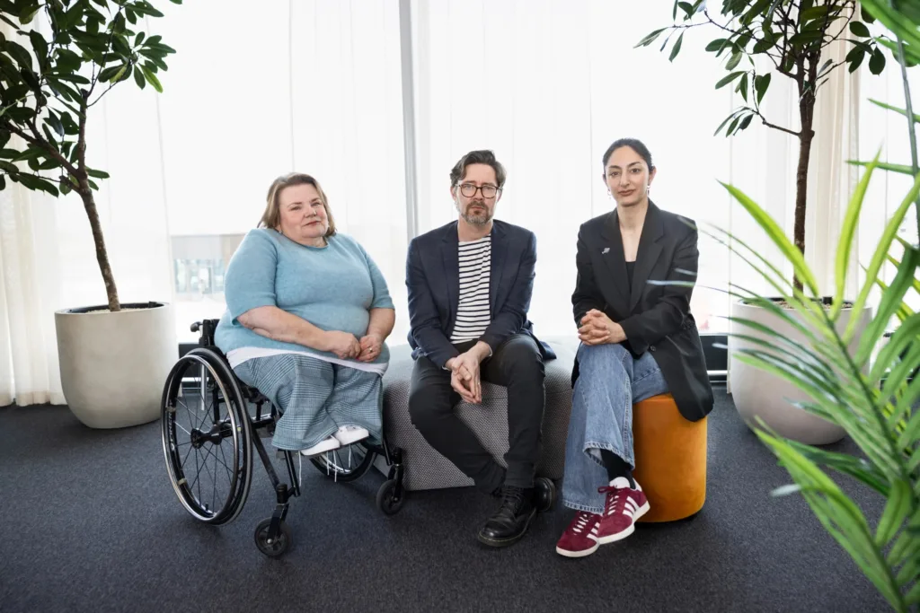 Samhalls Expertcentrum består av tre personer. På bilden sitter de tillsammans. Till vänster Maria Johansson, specialist funktionsrätt, i mitten Erik Hedin, specialist digital tillgänglighet och till höger Seher Yilmas, specialist i jämlikhet och inkludering. Maria sitter i en rullstol och har en ljusblå tröja. Erik sitter på en grå pall, har glasögon och är klädd i mörk kavaj. Seher sitter på en orange sittkudde och har också kavaj. Alla tre ser ganska allvarliga ut och tittar rakt in i kameran.