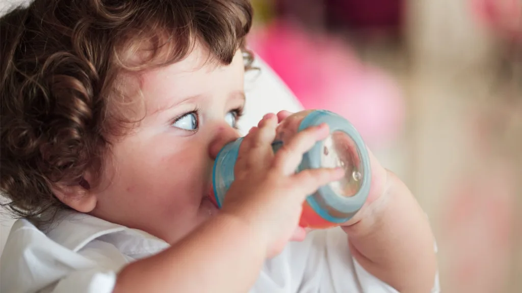Ett litet barn med lockigt hår. Hen dricker ur sin pipflaska.