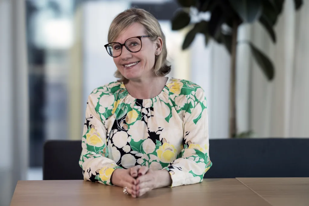 Maria Made sitter vid ett bord. Hon har ljust hår, mörka glasögonbågar och en mönstrad blus. Hon ler och tittar in i kameran. 