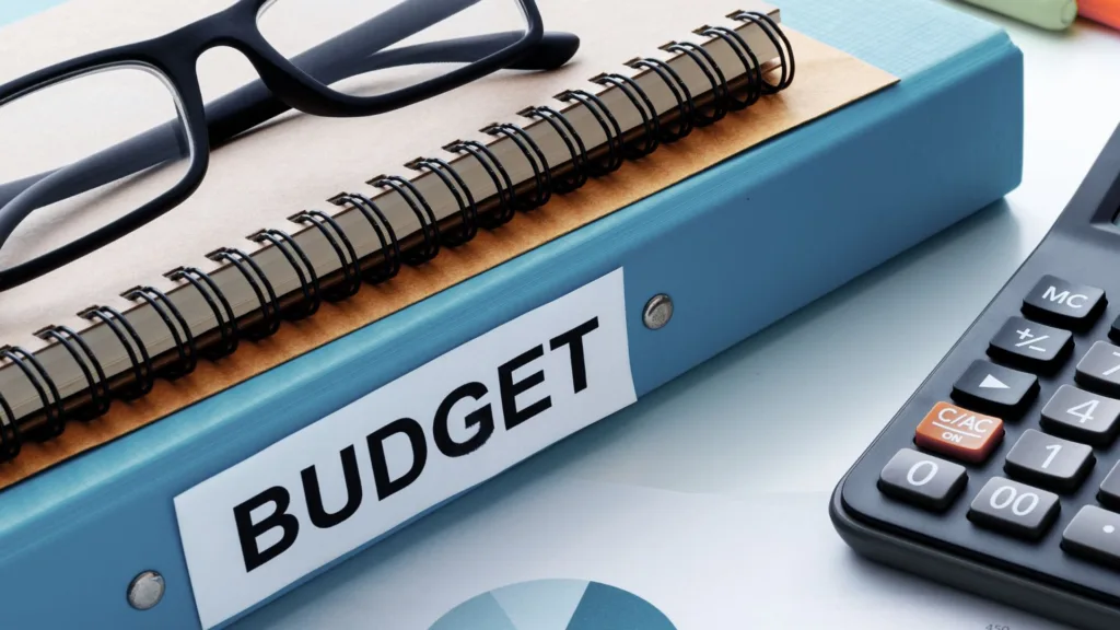 Miniräknare, block, läsglasögon och en pärm med texten "Budget" på ryggen.