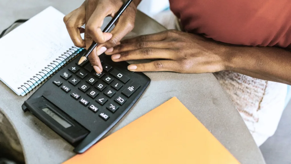 Kvinna sitter vid ett bord med miniräknare, penna och papper. Hennes händer i bild.