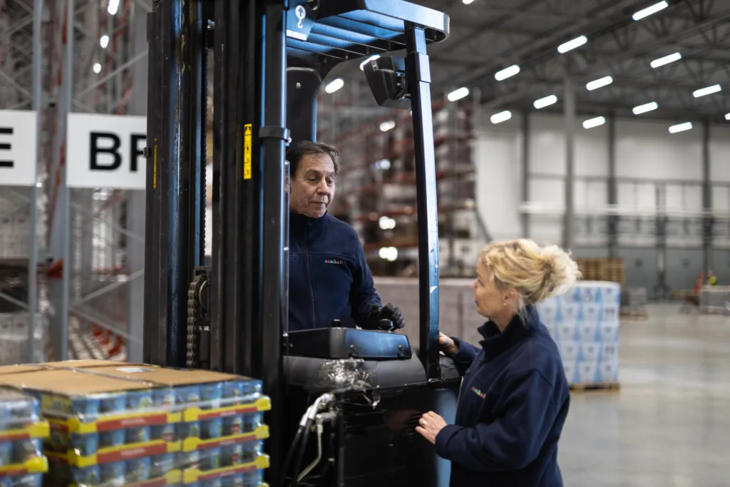 En kvinna står och pratar med en man som kör truck. Båda bär mörka Samhallkläder.